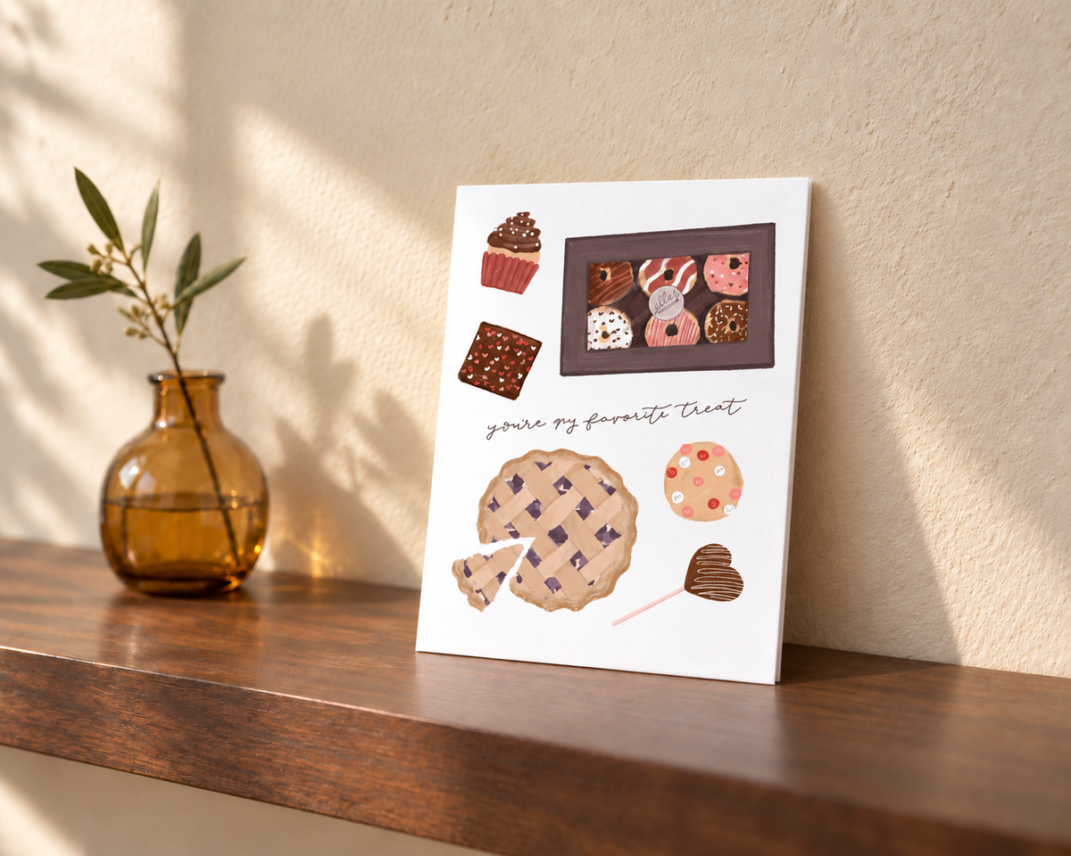 A white greeting card featuring watercolor illustrations of various desserts, including a cupcake, chocolate bar, donut, and a large lattice cookie, leaning on a wooden shelf next to an amber glass vase with a green stem.